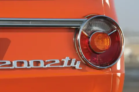 Detalle de la parte trasera de un BMW 2002 Tii naranja, con su icónico emblema cromado y una luz trasera circular.