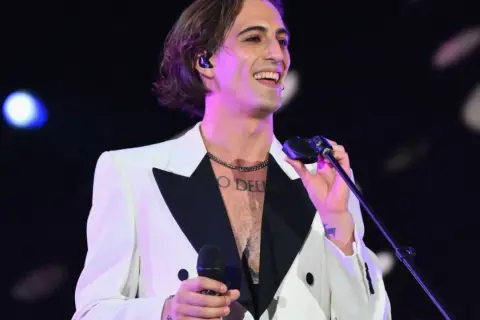 Damiano David w białej marynarce z czarnym kołnierzem śpiewa na koncercie, trzymając mikrofon.