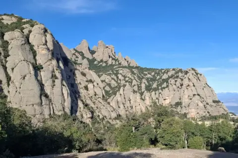 Imponentes rocas de Montserrat bajo un cielo azul, con vegetación verde y la creu de Sant Miquel en la distancia.