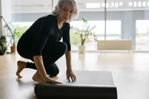 Une femme prépare son tapis pour faire du sport chez soi. Elle est à genoux, déroulant le tapis gris sur le sol en bois.