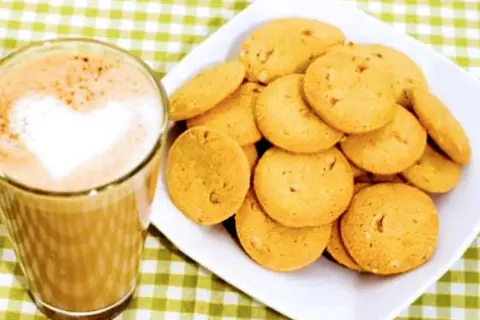 A glass of coffee with a heart-shaped foam art next to a plate of golden brown, round cookies.