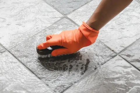 Mano con guante naranja cepillando un suelo antideslizante con espuma.