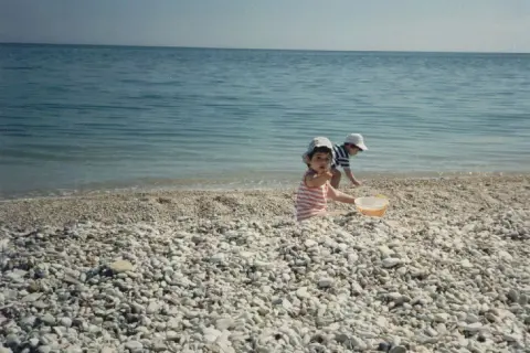 Bambini giocano sulla spiaggia di ciottoli a Marina di Montemarciano, con il mare azzurro sullo sfondo.