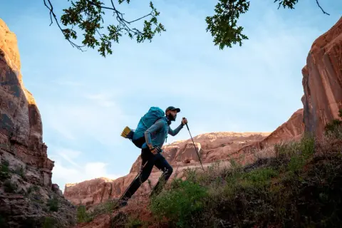 Hombre con mochila y bastones de senderismo sube una ladera rocosa bajo un cielo azul.