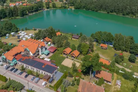 Widok z lotu ptaka na ośrodek wypoczynkowy nad turkusowym jeziorem. Widać budynki, domki, pole namiotowe, kampery i plażę. To idealne miejsce na wypoczynek, niczym złota łania wiartel.