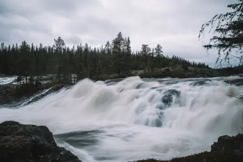 Vilt och mäktigt vattenfall i Norrland, med tät barrskog som bakgrund och en dramatisk, grå himmel.