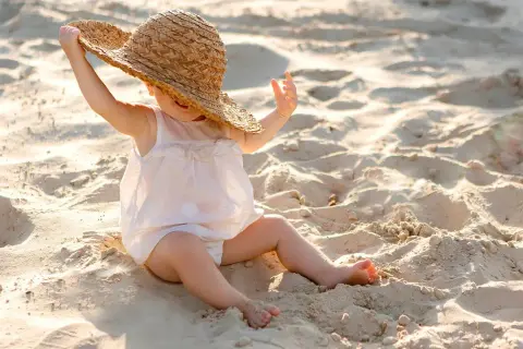 Liten bebis på stranden med solskyddsfaktor, leker med en stor stråhatt.