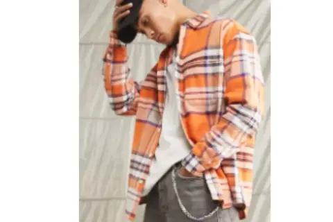 Uomo con camicia in flanella a quadri arancioni, jeans grigi e cappellino nero.