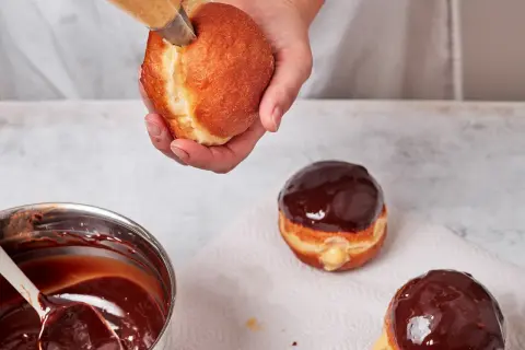 Eine Hand füllt einen Boston Cream Donut mit cremiger Füllung. Daneben liegen bereits glasierte Donuts.