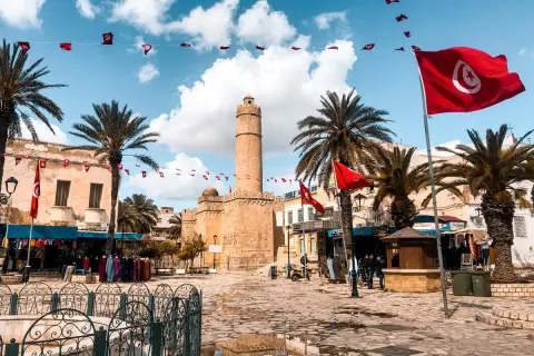 Placez-vous ici pour savoir où aller en Tunisie : une place animée avec des drapeaux tunisiens, des palmiers et une tour historique.