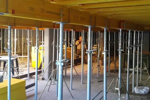 Metalowe stemple budowlane podtrzymują żółte belki stropowe. W tle widać pracownika i inne elementy konstrukcyjne.