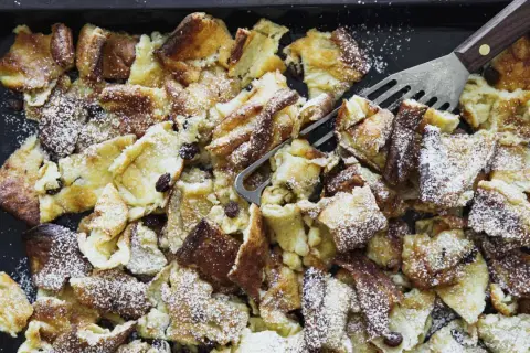 Kaiserschmarrn aus dem Backofen auf einem Blech, mit Puderzucker bestäubt und Rosinen. Ein Gabel-Schaber liegt bereit.