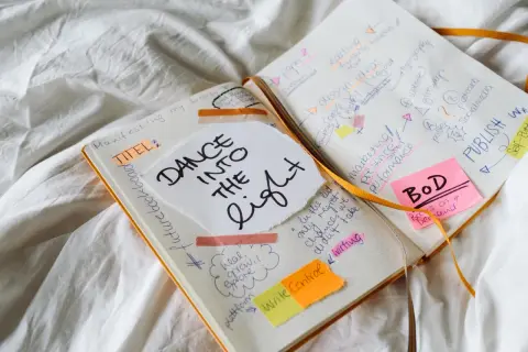 Un carnet ouvert avec des notes manuscrites sur la création d'un livre, comme "DANCE INTO THE LIGHT". On y voit des idées de titres, de marketing, et la question "a quoi sert un pseudo" est implicite dans la planification.