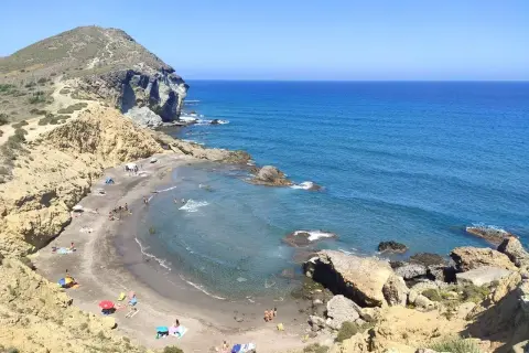 Vista aérea de **playas nudistas Cabo de Gata**. Arena oscura, aguas turquesas y formaciones rocosas. Gente disfrutando del sol y el mar.