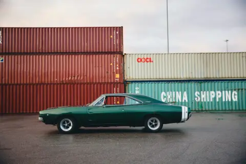 Klasyczny zielony **hatchback** Dodge Charger z białym pasem na tyle, zaparkowany przy stosach kontenerów w porcie.