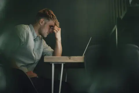 Ein Mann sitzt mit der Hand an der Stirn vor einem Laptop. Er wirkt nachdenklich, aber es ist möglich, dass er trotz Depressionen arbeitet.