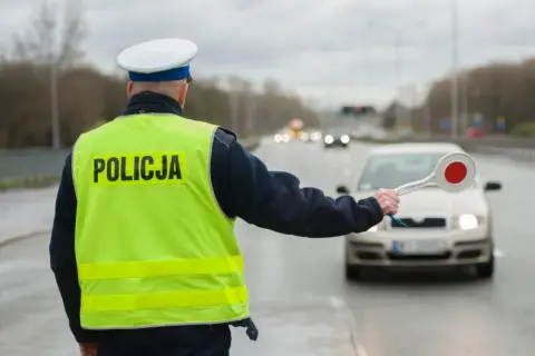 Policjant w kamizelce odblaskowej zatrzymuje samochód. Kierowca rozmawiający przez telefon może spodziewać się mandatu.