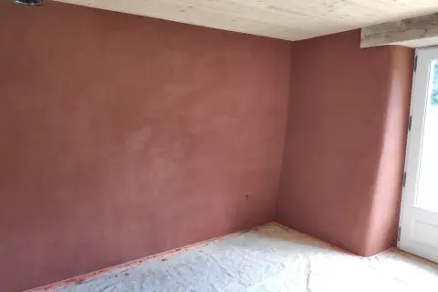 Un mur d'angle avec un enduit chaux sable intérieur d'une belle teinte ocre, sous un plafond en bois clair. Le sol est protégé par une bâche.