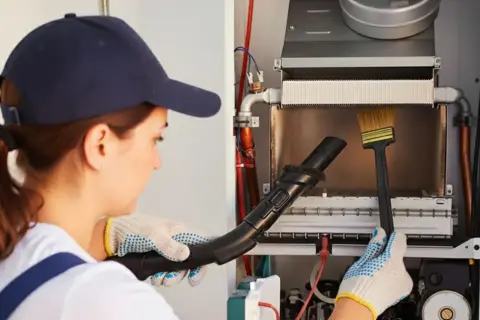 Technicienne effectuant le ramonage d'une chaudière à condensation avec un aspirateur et une brosse.