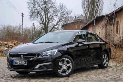 Czarny Peugeot 508 2.0 HDI na brukowanej drodze, w tle zniszczone budynki i drzewa.