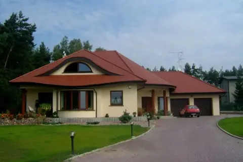 Elegancki dom w stylu włoskim z czerwoną dachówką, otoczony zielenią. Na podjeździe stoi czerwony samochód.