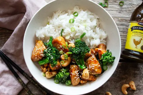 En skål med ris, kyckling och broccoli, toppad med sesamfrön och salladslök. Bredvid syns en flaska kycklingfond.