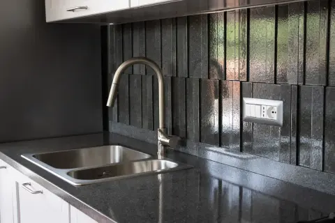 Top cucina in pietra lavica: pro e contro. Un lavello doppio in acciaio inox e un rubinetto moderno su un piano scuro e un rivestimento a listelli verticali.
