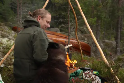 En man med en hund sitter vid en lägereld i skogen. En kanot ligger i bakgrunden. Detta är vad en fotosession betyder för dem.
