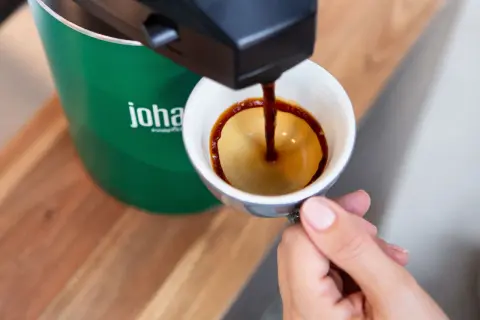 En hand håller en kopp medan kaffe rinner ner från en maskin. Frågan om hur många matskedar kaffe per kopp är relevant för att få den perfekta smaken.