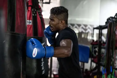 Boxer in blue gloves throwing a powerful cross punch at a heavy bag, sweat glistening on his arms.