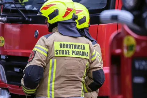 Strażacy w żółtych hełmach i beżowych kurtkach z napisem "Ochotnicza Straż Pożarna" stoją obok czerwonego wozu strażackiego.