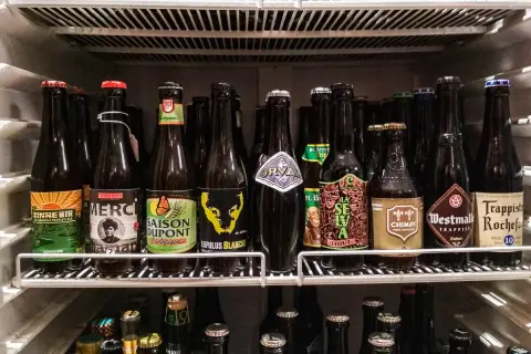 Scaffale pieno di bottiglie di birra, alcune con etichette colorate come "Zinnebir" e "Saison Dupont". Una birra aperta in frigo, pronta per essere gustata.