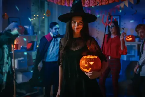 Kiedy obchodzi się Halloween, młoda kobieta w stroju czarownicy trzyma świecącą dynię na imprezie z przyjaciółmi w kostiumach.