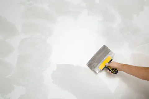 Mano con espátula amarilla y gris trabajando en una pared, alisando la textura para como quitar gotele.