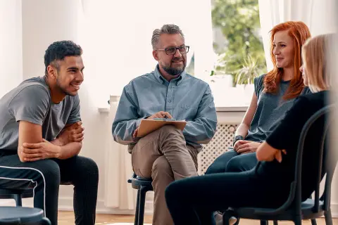 Grupa ludzi na sesji terapeutycznej. Mężczyzna z notatkami prowadzi rozmowę, a uczestnicy słuchają. Może uczą się języka francuskiego?