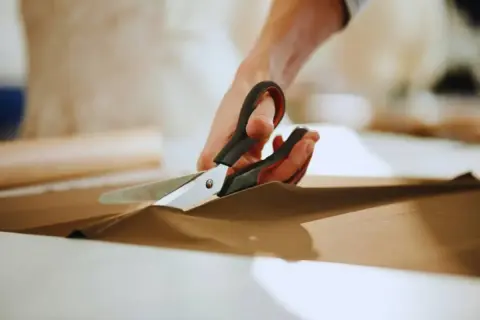 Ręce trzymają nożyce, precyzyjnie wykonując cięcie na brązowym materiale. To przykład podstawowych technik krojenia.