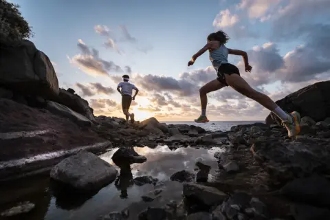 Deux coureurs s'entraînent sur un sentier rocailleux à Tenerife au coucher du soleil, l'océan en arrière-plan.
