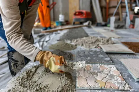 Un ouvrier applique du mortier pour un ragréage de sol. Des tas de sable et de ciment sont prêts pour le travail.