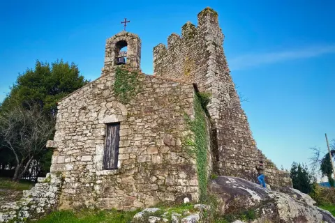 Antigua ermita de piedra con campanario y cruz, rodeada de vegetación. Las **torres de oeste** se alzan imponentes contra el cielo azul.