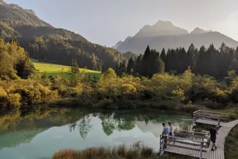 Słowenia gdzie na wakacje? Jezioro o turkusowej wodzie, otoczone jesiennymi drzewami i górami. Ludzie spacerują po drewnianych pomostach.
