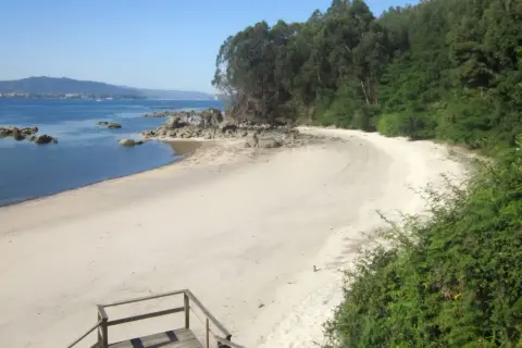Escaleras de madera bajan a la playa Niño do Corvo, con arena blanca, rocas y mar azul.
