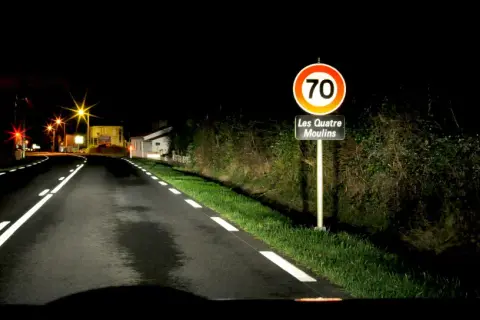 Route de nuit avec marquage au sol blanc lumineux. Panneau "Les Quatre Moulins" et limite de vitesse à 70.