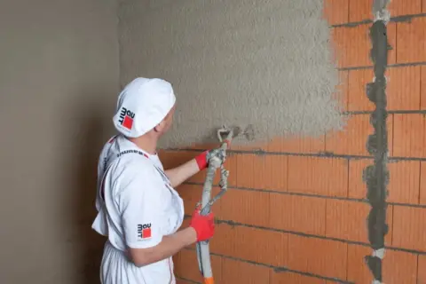 Pracownik w białym ubraniu i czapce z logo Baumit nakłada tynk agregatem na ścianę z pustaków. Obliczasz ile kg tynku na m2?