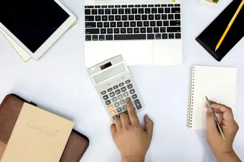 Ręka naciska przycisk kalkulatora, by sprawdzić, co to jest saldo rachunku. Obok leży laptop, tablet i notatnik.