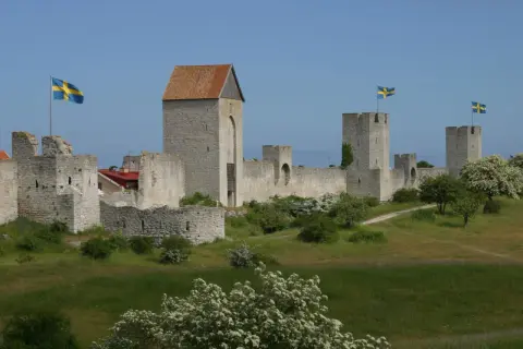 Visby muren reser sig stolt mot en blå himmel, med svenska flaggor som vajar från tornen. En historisk stadsvall som vittnar om svunna tider.