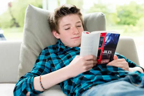 Un adolescent aux taches de rousseur lit un livre captivant, plongé dans un monde d'aventures. Il est confortablement installé sur un canapé, entouré de livres pour adolescents.