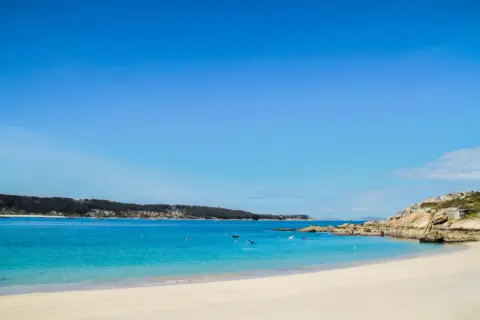 Arena blanca y aguas turquesas en la playa de Menduiña, con barcas a lo lejos y una costa rocosa.