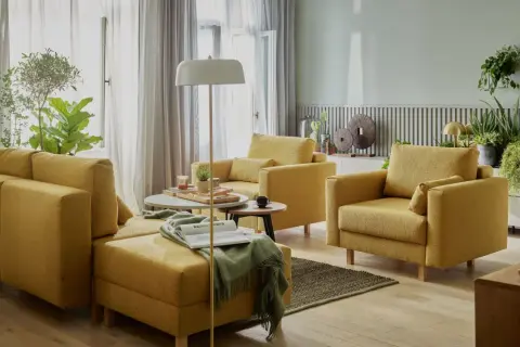 Przytulny salon bez telewizora. Żółte meble wypoczynkowe, stoliki kawowe, lampa podłogowa i mnóstwo zielonych roślin tworzą relaksującą atmosferę.