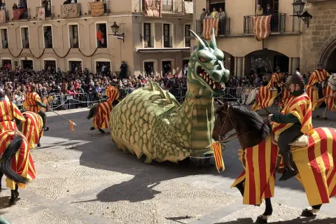 Un dragón gigante desfila en las fiestas de Alcañiz, acompañado por jinetes con trajes de rayas rojas y amarillas.