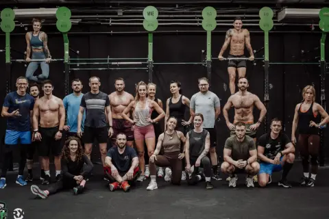Grupa osób w strojach sportowych pozuje na tle sali treningowej CrossFit.